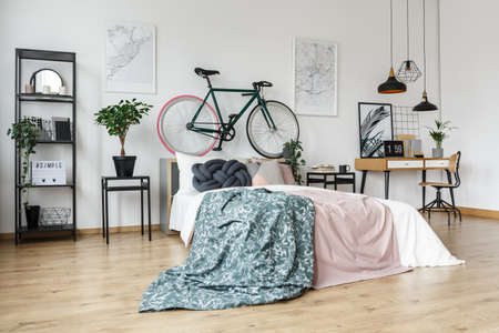 Side angle of pastel bed with green blanket and bike next to simple study space with laptop on...