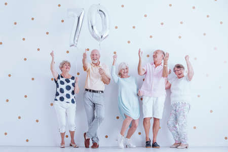 Smiling grandparents enjoying friend's birthday. Couple holding silver balloons against white wall with gold dotsの写真素材