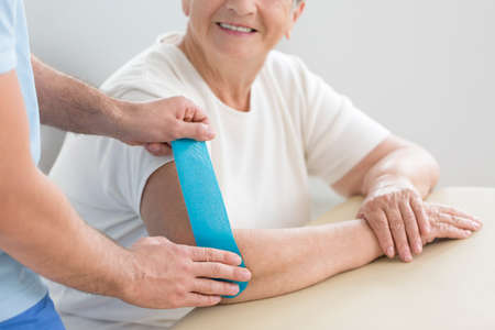 Close-up of physiotherapist putting tape on arm of smiling senior woman during kinesiotaping therapyの写真素材