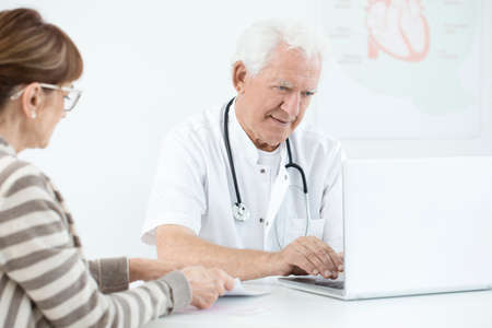 Elderly doctor using laptop during medical consultation with patient in white officeの写真素材