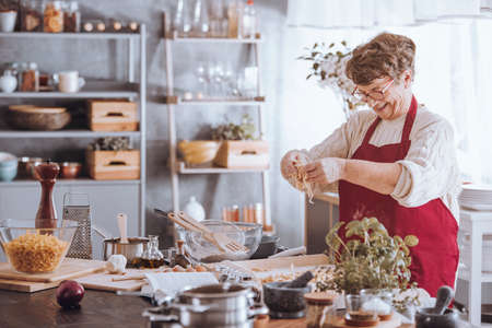 Grandmother adding egg to flour while cooking in the kitchenの写真素材