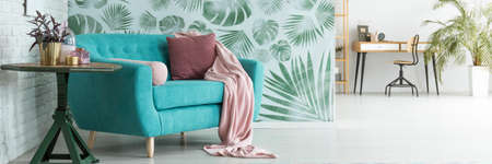 Pink blanket and pillow on turquoise sofa next to table with plant in gold pot against floral...