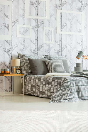 Table with clock, lamp and decorations standing by the bed with patterned grey and white bedclothes...