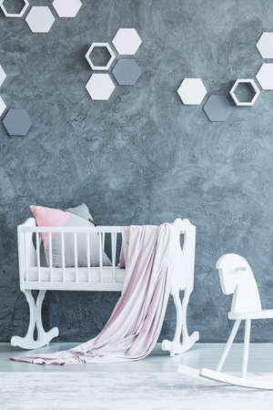 Pastel blanket on baby's crib next to a rocking horse in bedroom interior with white honeycombs on concrete wallの写真素材