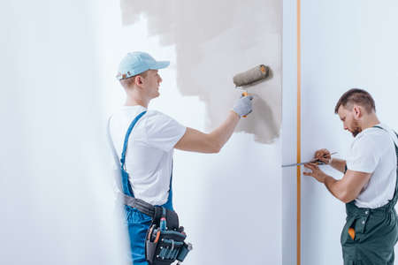 Painter with tool belt painting the wall and his colleague measuring the wall during finishing interiorの写真素材