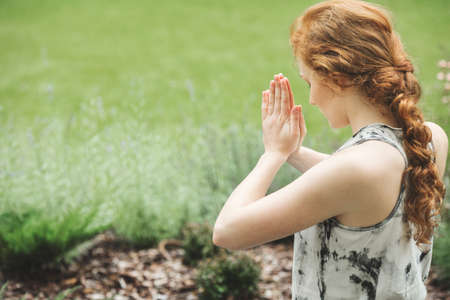 Focused red-haired woman with joined hands meditating in the garden