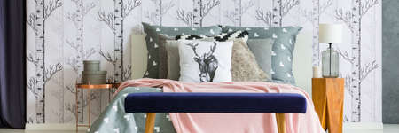 Dark blue bench in front of king-size bed with deer pillow in bedroom with lamp on wooden stoolの写真素材