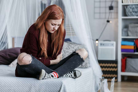 Red-haired, young girl sitting on her bed with stomach pain