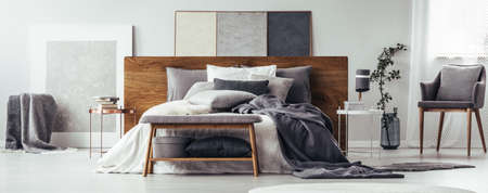Books on copper table and bench next to bed with wooden bedhead in modern bedroom interior with grey chairの写真素材