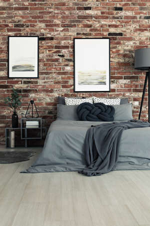 Dark bedding on king-size bed in bedroom with gallery on brick wall and vase on nightstandの写真素材