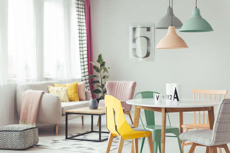 Pastel lamps above wooden round table in bright living room interior with patterned pouf and yellow cushion on a sofaの写真素材