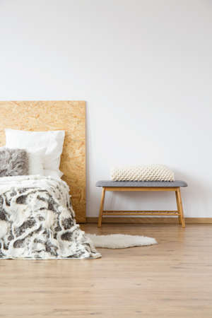 White knit blanket on bench next to bed with patterned bedsheets in natural bedroom with copy space