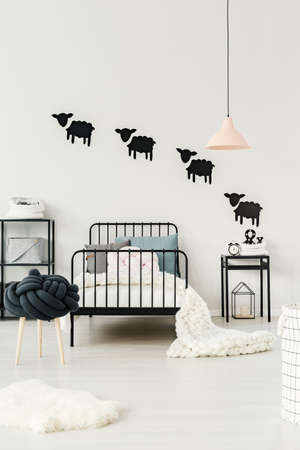 White fur next to a stool and kid's bed with blanket under pastel lamp in bedroom interior with...