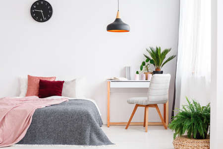 Grey chair at desk with plant against white wall with clock in pink bedroom interior with bed, lamp and fernの写真素材