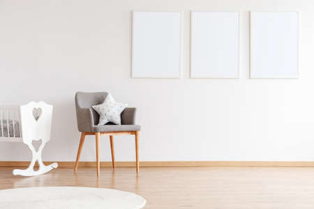Empty posters hanging on white wall in monochromatic nursery interior with gray armchair and white...