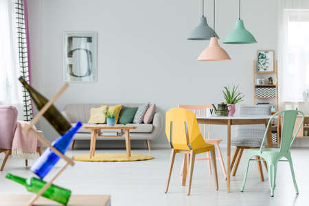 Mint and yellow chair at table with kettle under pastel lamps in spacious flat interior with cushions on sofa near yellow round carpet