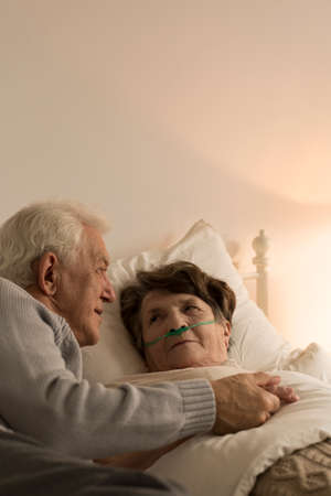 Elderly man taking care of his sickly wife who is lying in bed being lit by a dim lamp light