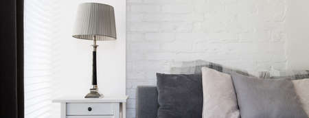 Close-up of nightstand with lamp near the bed with grey pillows on a background of white brick wall
