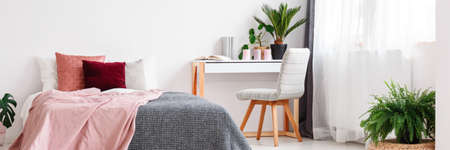 Pink and grey bedsheets on bed in cozy bedroom interior with chair at desk with plants, candles and booksの写真素材