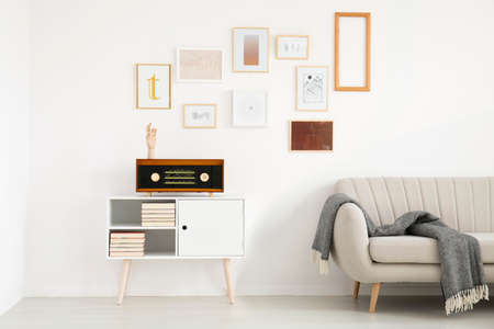 Blanket on beige sofa next to white cupboard with radio against the wall with mockup of poster in simple living room interiorの写真素材