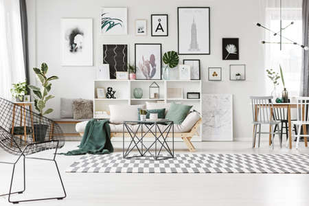 Metal chair and table in bright living room interior with green pillows on the couch, against a wall with gallery of postersの写真素材
