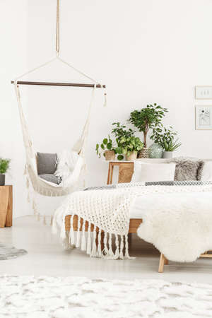 Cozy bedroom interior with beige blanket and fur on wooden bed and gray cushion on a hammock