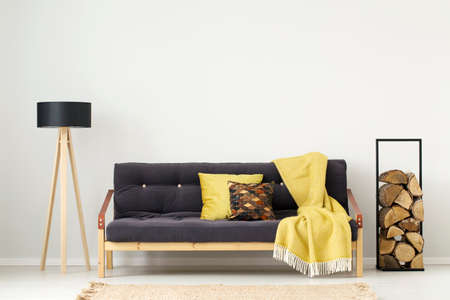 Black sofa with yellow blanket between logs of wood and lamp in apartment interior with copy spaceの写真素材