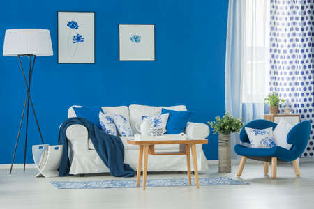 Patterned pillows on white couch in blue living room interior with flowers posters and wooden tableの写真素材
