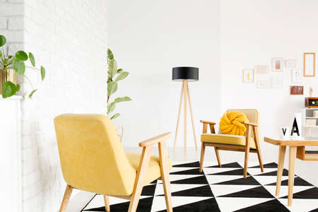Yellow armchairs and table on black and white carpet in bright flat interior with wooden lamp