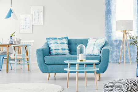 Candle on blue round table in front of a sofa with patterned cushions in flat interior with wooden lamp and posters on white wallのeditorial素材