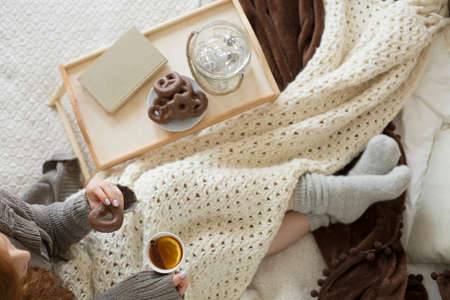 High angle of woman drinking tea with lemon while lying in bed with pretzels on wooden tray