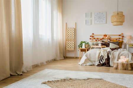 Candles on wooden stump next to bed with knit blanket in hygge style bedroom interior with brown curtain and postersの写真素材