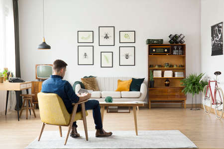 Man sitting in yellow chair at wooden table in retro living room interior with sideboard and gallery above a setteeの写真素材