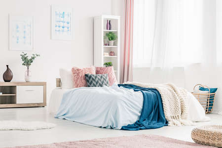 Pink pillows and blue blanket on bed in womanly bedroom interior with plant on wooden cupboardの写真素材