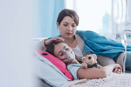 Worried mother taking care of a kid with cystic fibrosis lying in hospital bed with plush toyの写真素材