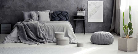 Cactus, pouf and boxes near bed in grey bedroom interior with bright poster on the wall
