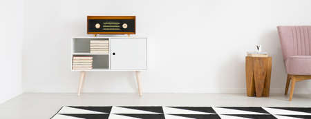 Radio on white cupboard near wooden stool and pink chair in bright living room interior with geometric carpetの写真素材