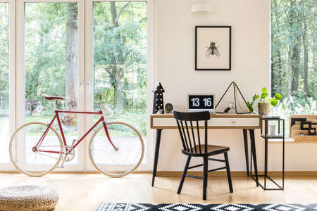Black chair at wooden desk with laptop and lamp in workspace with poster and red bicycleの写真素材