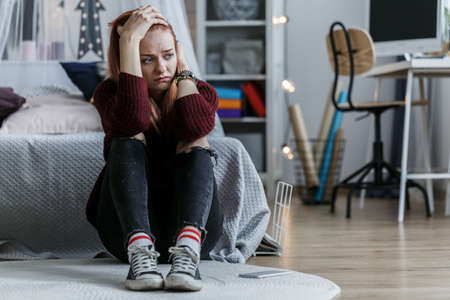 Worried, young girl holding her head, witting on the floor in her bedroom