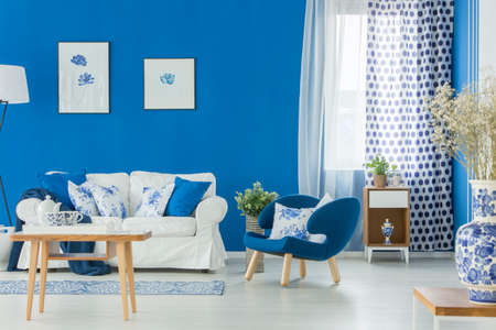 Blue armchair and white couch in living room interior with flowers, posters and wooden table