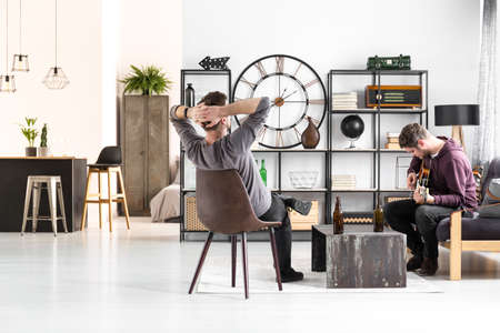 Two men spending time together playing the guitar and drinking beer in open space apartment interiorの写真素材
