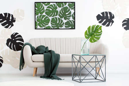 Metal table in front of sofa with dark blanket in floral living room interior with monstera leaves...