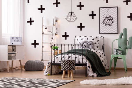 Patterned wallpaper, stool and carpet in kid's bedroom interior with cactus pillow on green chair next to bedの写真素材