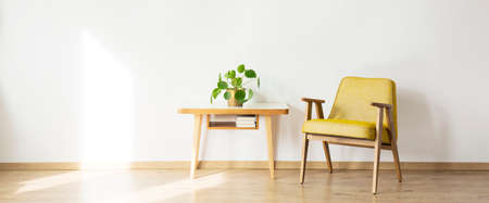 Vintage yellow armchair standing next to a wooden table with plant on it in bright waiting roomの写真素材