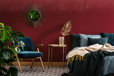 Gold table between green armchair and bed in sophisticated red bedroom interior with mirrorの写真素材