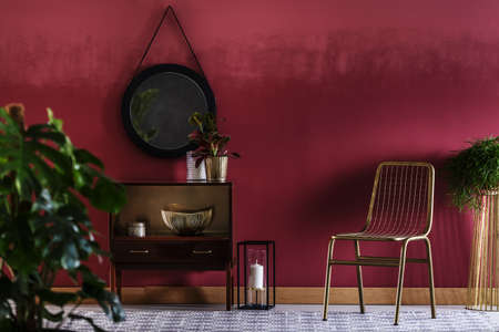 Gold chair next to a cabinet with plant against red wall with mirror in living room interior with a candleの写真素材