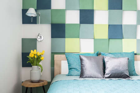 Colorful wall in bedroom interior with silver pillows on turquoise bed next to stool with tulipsの写真素材