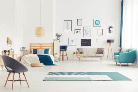 Blue armchair near beige sofa and lamp in cozy open space interior with bed and postersの写真素材