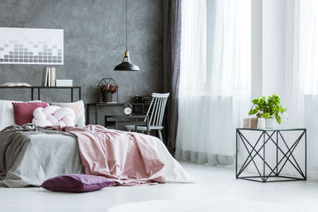 Metal table next to the window in bright bedroom interior with pink bed against gray, concrete wallの写真素材