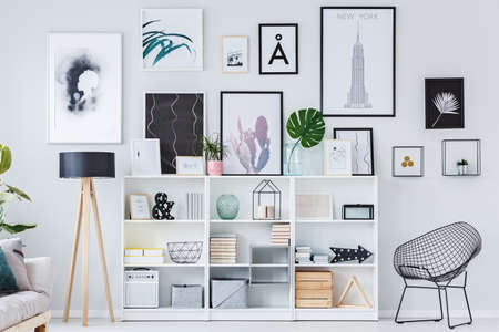 Wooden shelves with decorations, boxes and books in white living room interior with fresh plants and modern art postersの写真素材